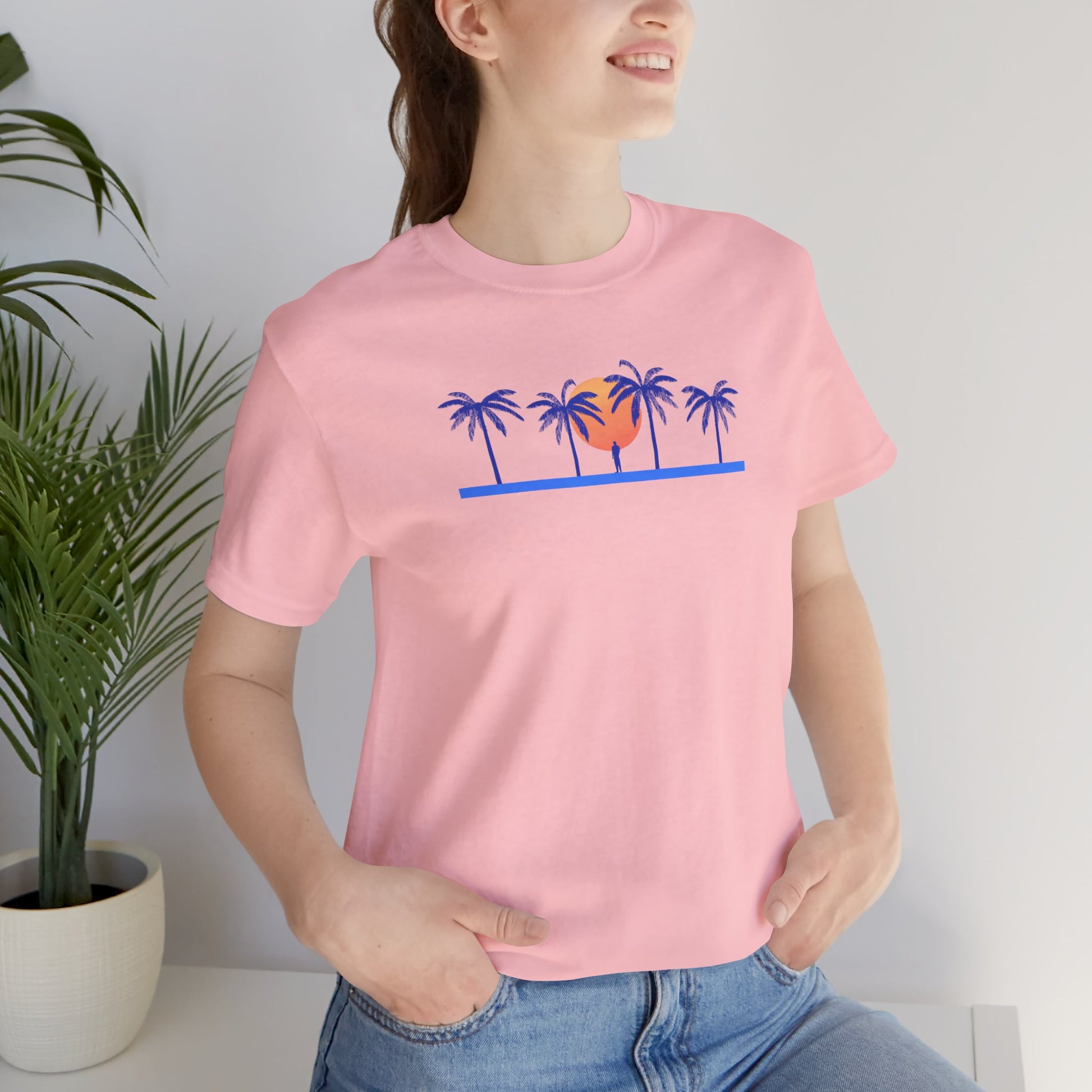 Retro unisex tropical pink t-shirt with palm trees and sunset illustration, worn by a woman